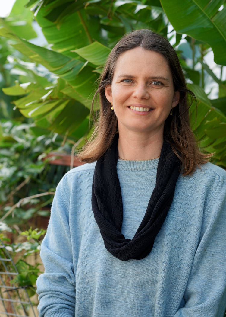 Woman wearing a blue sweater and black scarf standing in front of green foliage