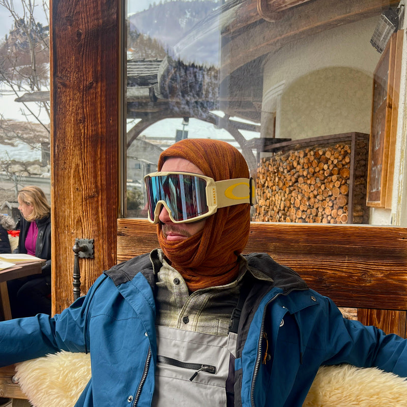 Person wearing ski goggles and a blue jacket in a cozy indoor setting with wooden walls and a mountain view. While wearing a redwood merino beanie wrap as a snood/hood.